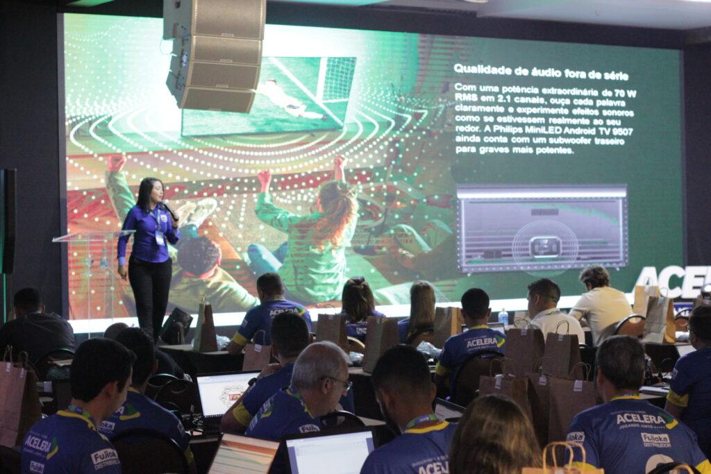 Palestra do evento Acelera. Pessoas sentadas em frente a notebooks enquanto assistem à uma palestrante. A palestrante está em cima do palco. Atrás há um painel de LED com o conteúdo da apresentação. Palestrante e convidados estão usando uma blusa personalizada do evento.